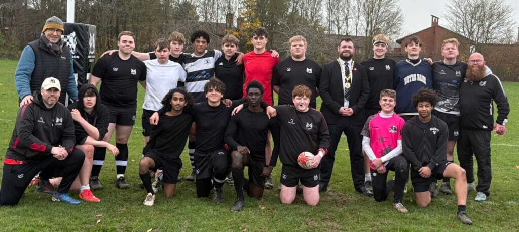 Group of rugby players and coaches posing on a grass field, wearing black jerseys, with some kneeling in front.