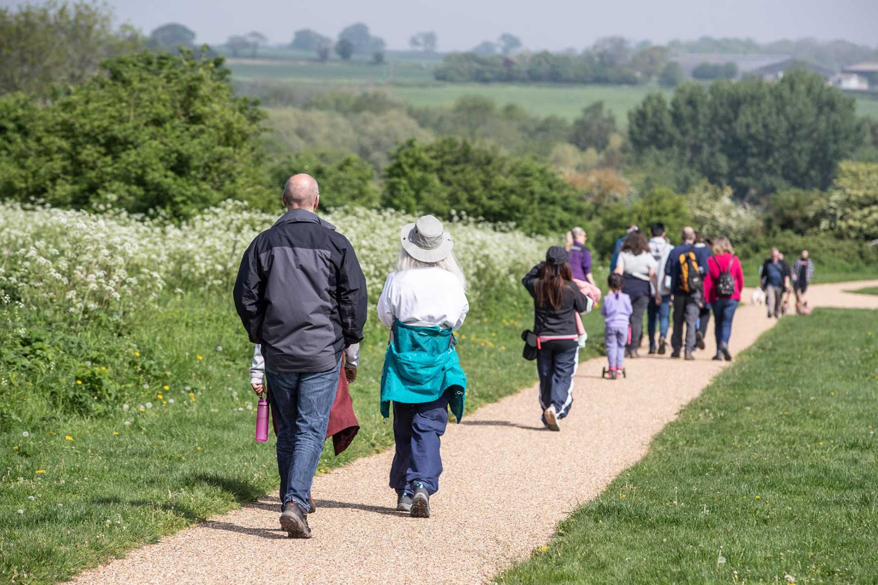 Group of people walking along a gravel path through a grassy meadow with trees in the distance; many wear backpacks and hats.