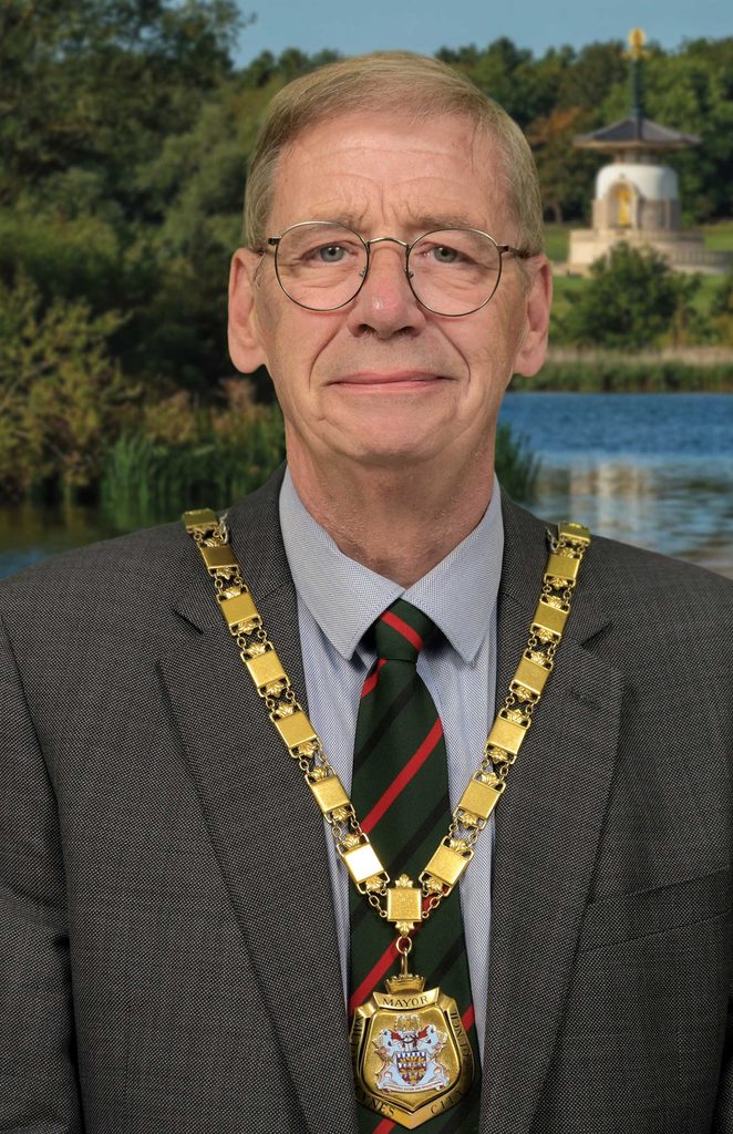 Portrait of a man in a suit wearing a gold mayoral chain, outdoors with trees and a pond behind him in a park.