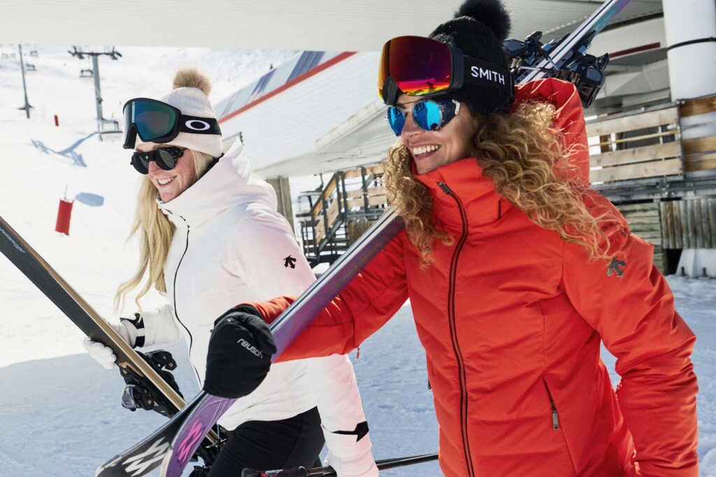 Ellis-Brigham-Female-Duo-Descente_MeribelSki2026_131-Licence-Expires-16-09-2028--copy | MK Pulse Two women in ski gear carry their skis on a snowy slope at a ski resort, smiling.