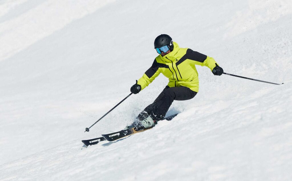 Ellis-Brigham_MeribelSki2026_158-Licence-Expires-16-09-2028--copy | MK Pulse Skier in a neon yellow jacket and black pants carving downhill in fresh powder, wearing a helmet and blue goggles.