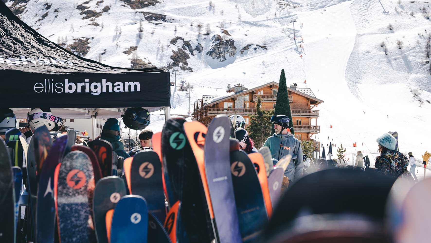Crowded ski resort scene under an Ellis Brigham tent with colorful snowboards in the foreground and a wooden lodge in the snowy background.